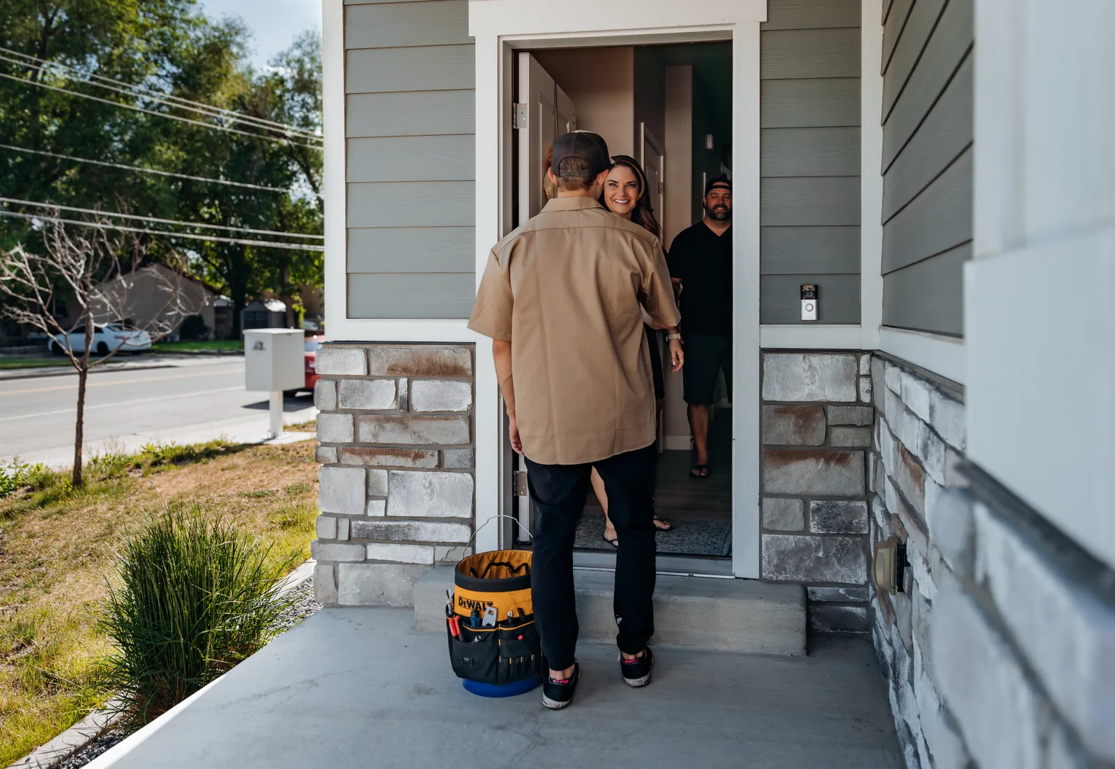 Blake from PowerClean greeting two smiling homeowners at the front door of a stone-and-siding home, his tool bag on the porch