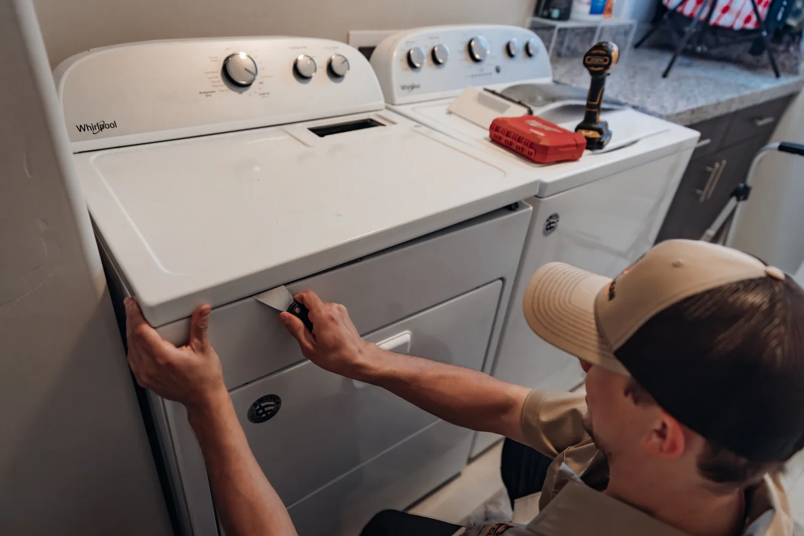 Blake from PowerClean sliding a putty knife into the seam under the top panel of a Whirlpool dryer, his other hand gripping the panel's corner