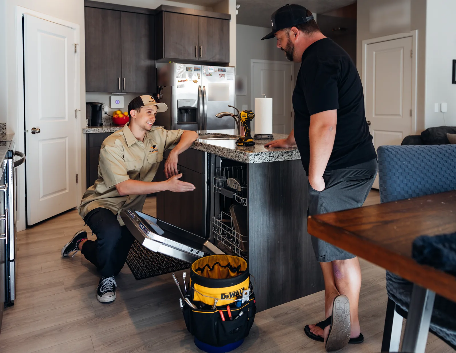 Blake from PowerClean kneeling beside an open dishwasher in a kitchen island, gesturing toward it as he smiles up at a customer standing at the end of the island