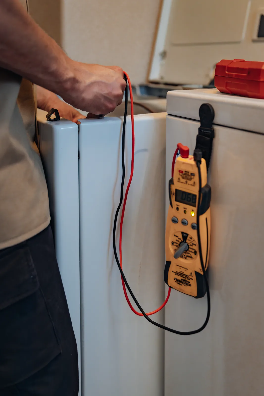 A Fieldpiece multimeter hanging by a strap from a washer, its red and black leads running up to Blake from PowerClean's hand at the dryer beside it