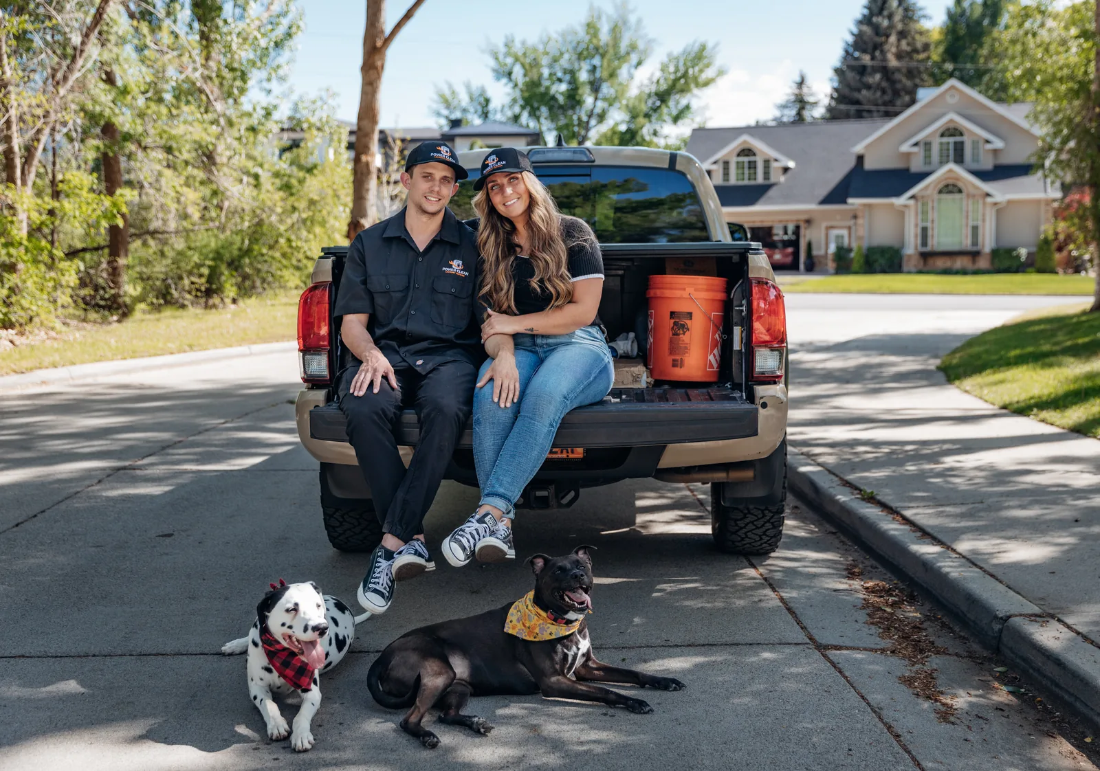 Blake Lonsberry and Makayla Adams of PowerClean on the tailgate of a tan pickup truck with two dogs, a Dalmatian and a black dog, in front of them