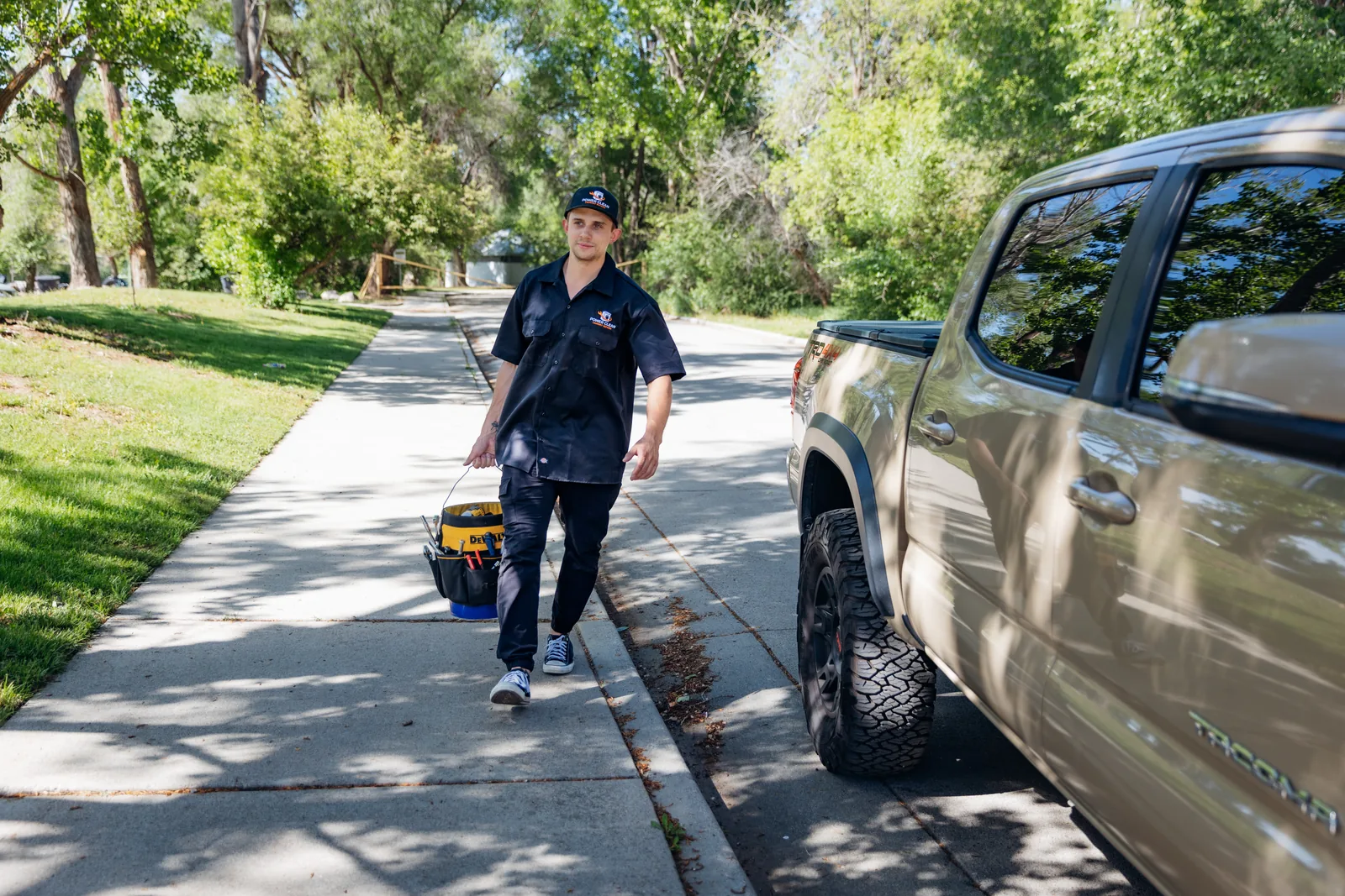 Blake from PowerClean walking toward the camera along a tree-lined sidewalk carrying a tool bag, a tan pickup truck parked beside him