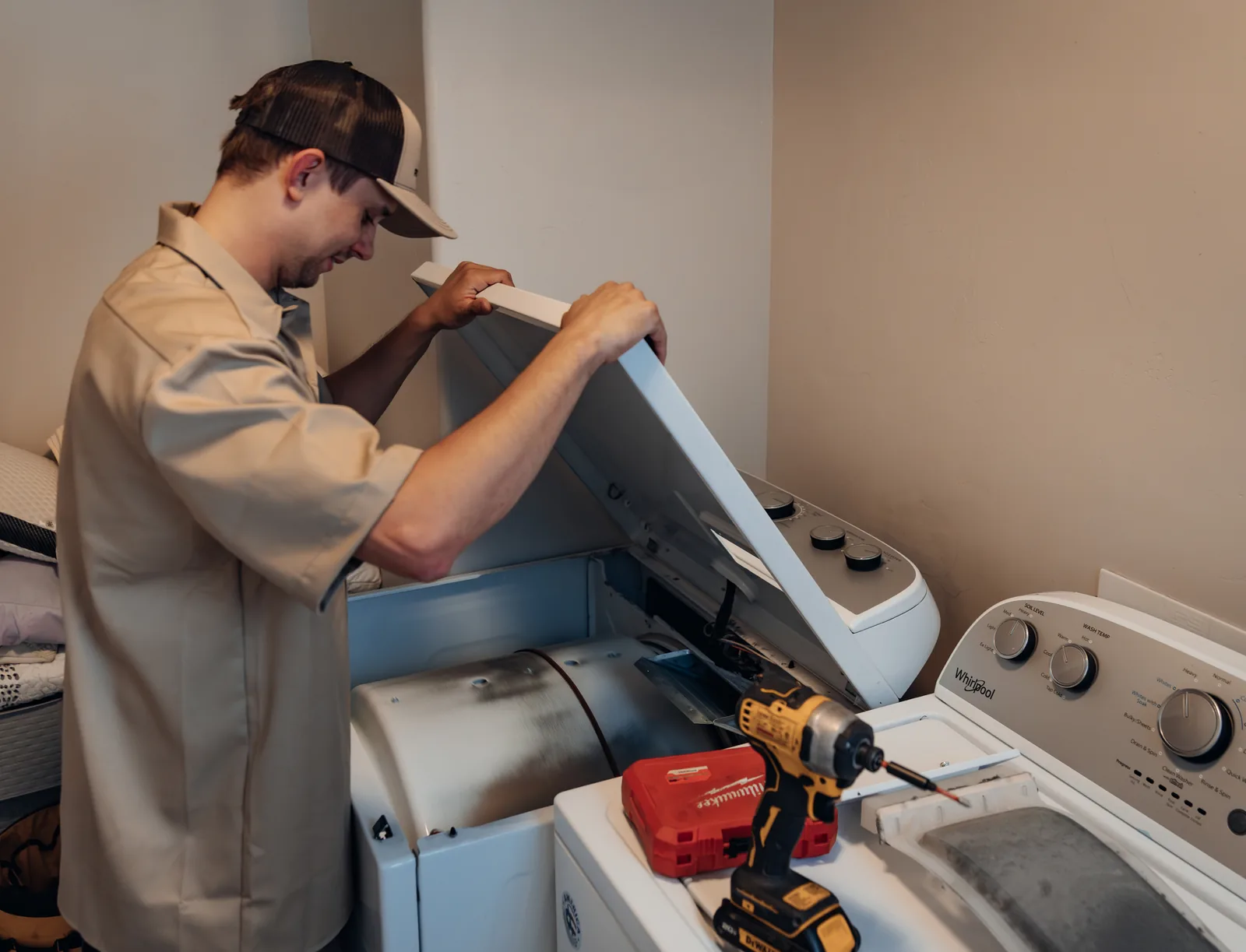 Blake from PowerClean raising a dryer's top panel, exposing the drum and drive belt, his impact driver and bit case on the Whirlpool washer beside it