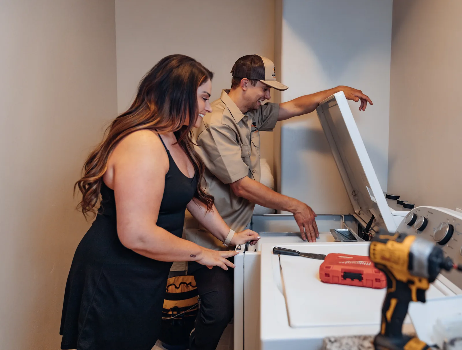 Blake from PowerClean reaching into a dryer, one arm over its raised top panel, beside a smiling customer, a bit case on the washer in front