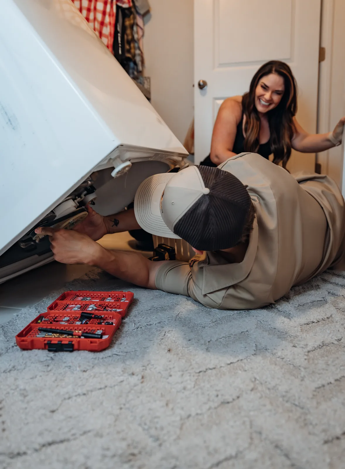 Blake from PowerClean reaching under a tilted-up washer with both hands while a customer crouched by the door behind him laughs